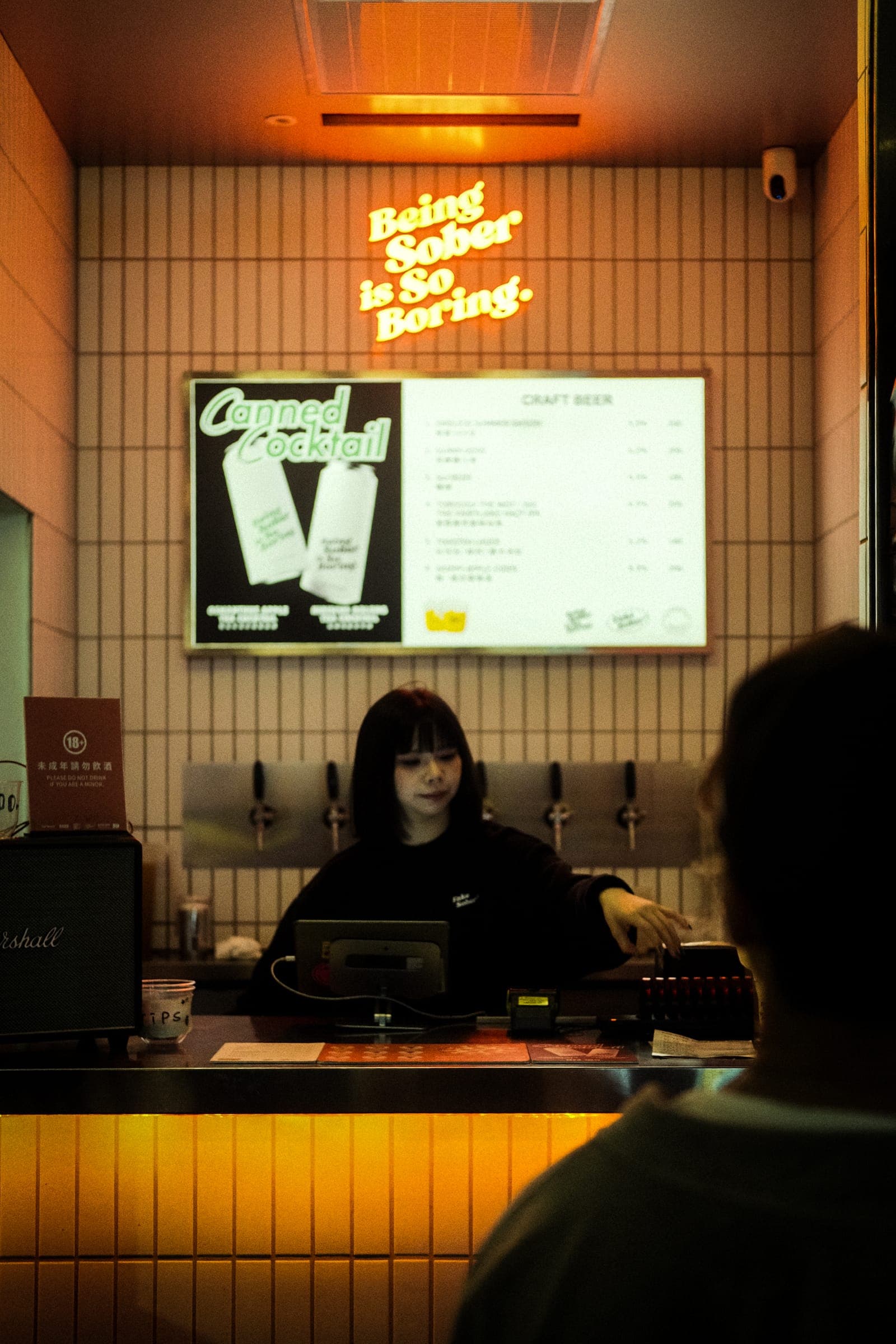 A bartender at a counter beneath a 'Being Sober is So Boring' neon sign.