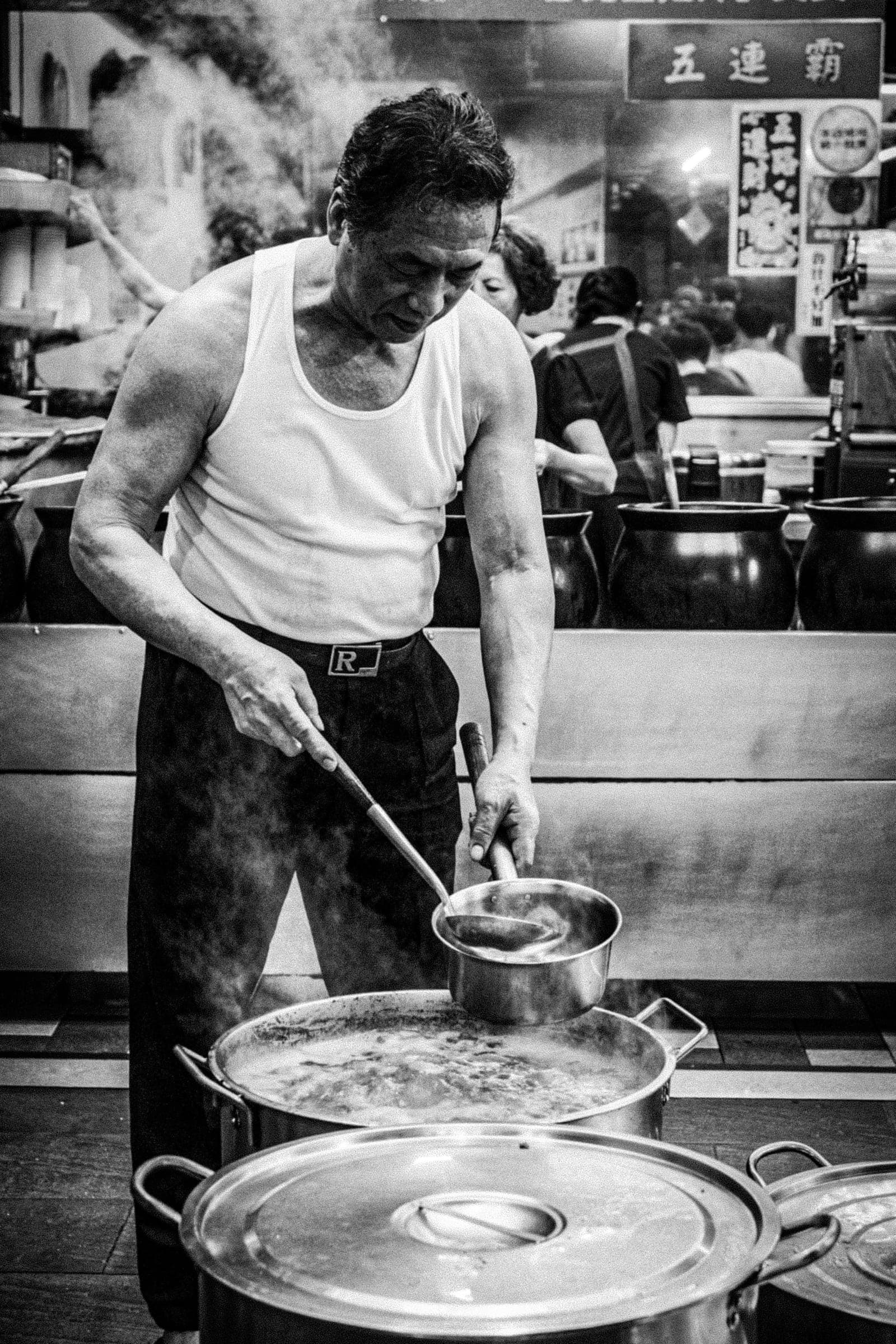 A cook in a white tank top stirring large pots in a kitchen filled with steam.