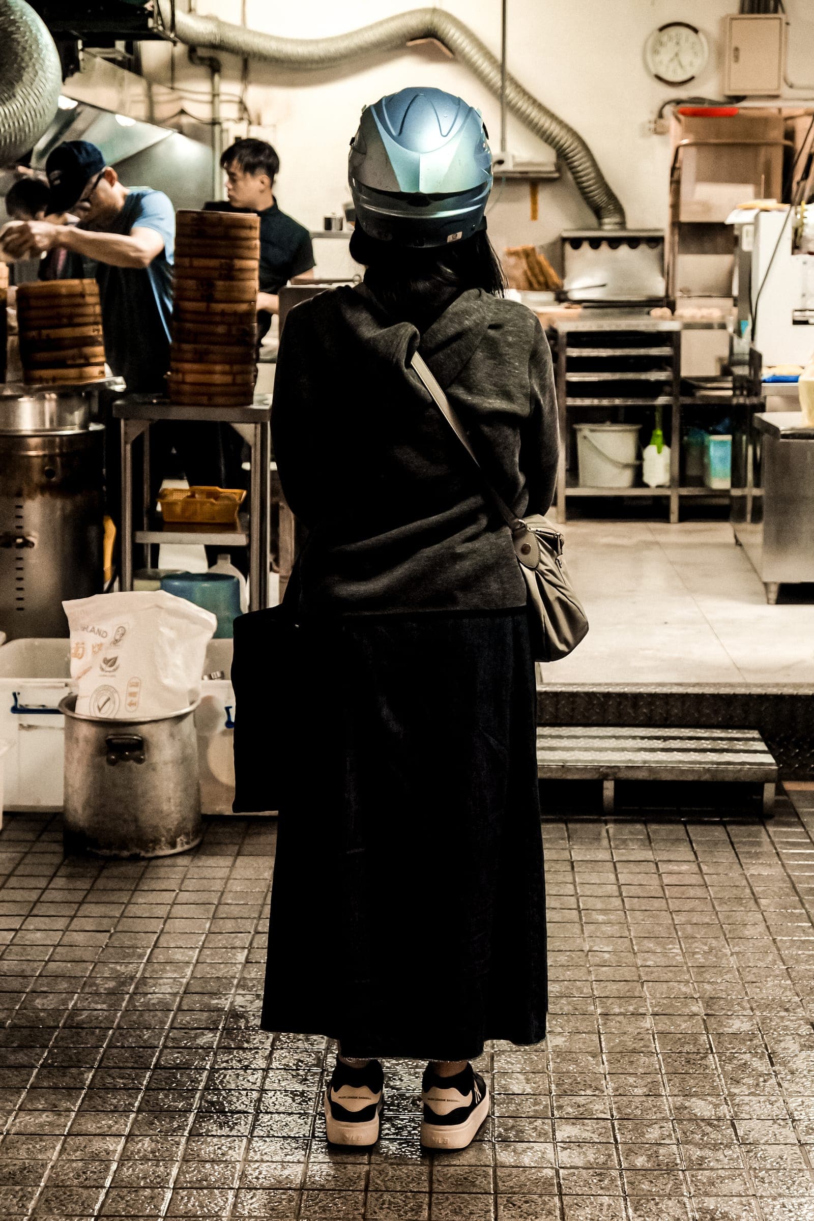 A figure in a long coat and motorcycle helmet standing in a steam-filled night-market kitchen.
